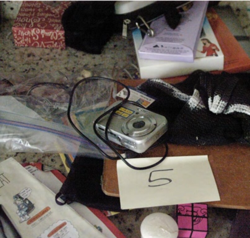FBI search photo of a digital camera labeled Item 5 on a cluttered countertop — absent from the entry photo of the same surface, and left behind at the scene